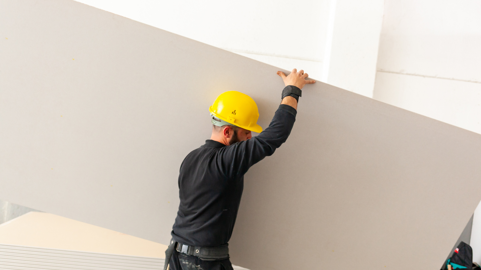 Un trabajador con casco amarillo y ropa de trabajo está montando un panel de yeso grande en una pared, en un entorno de construcción.