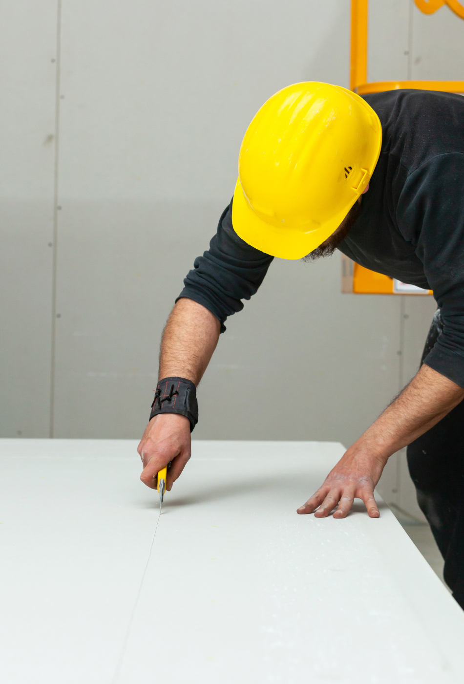 Un hombre en un sitio de construcción corta un panel de yeso con un cúter, usando guantes y casco amarillo, mostrando el proceso detallado de corte.